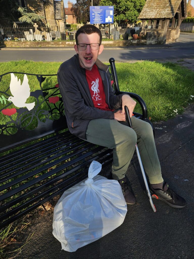 A 24-year-old man sits on a bench n the sunshine. He is holding a litter-picker and has a bag of rubbish at his feet. 